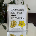 A clear acrylic coffee bag holder with a white paper insert displaying the brand 'SANDBOX COFFEE ROASTERS' and product details for 'red honey kurume dega', with illustrations of a melon and a yellow flower.