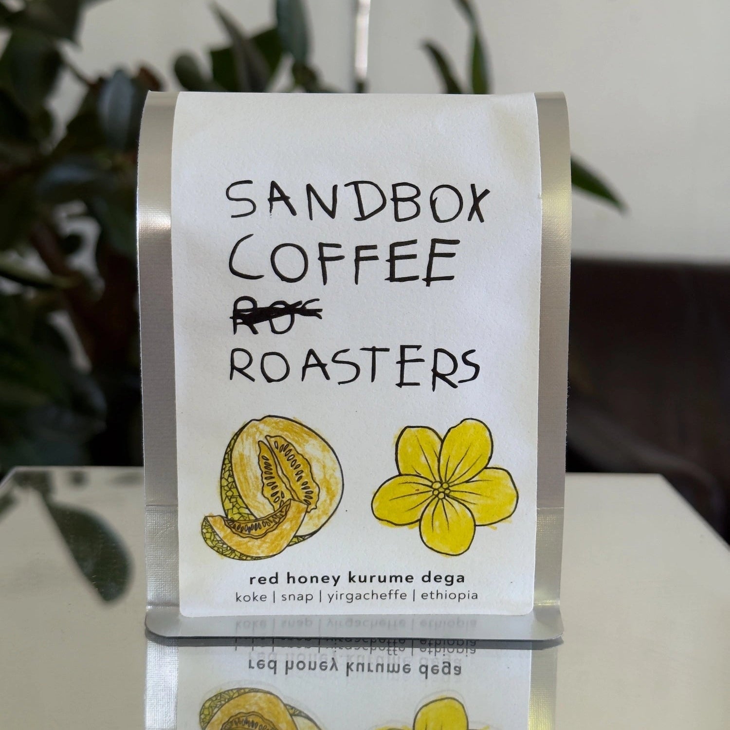 A clear acrylic coffee bag holder with a white paper insert displaying the brand 'SANDBOX COFFEE ROASTERS' and product details for 'red honey kurume dega', with illustrations of a melon and a yellow flower.